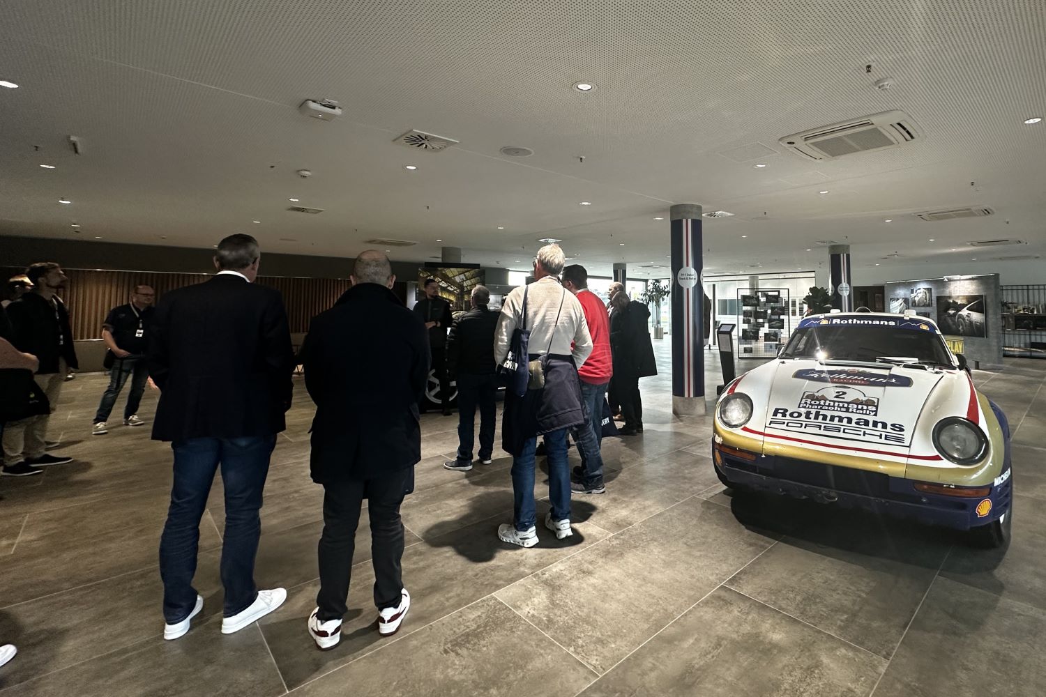 Personen, die im Kreis um einen Mann herumstehen und diesem zuhören. Im Vordergrund ein älteres Porsche Modell.