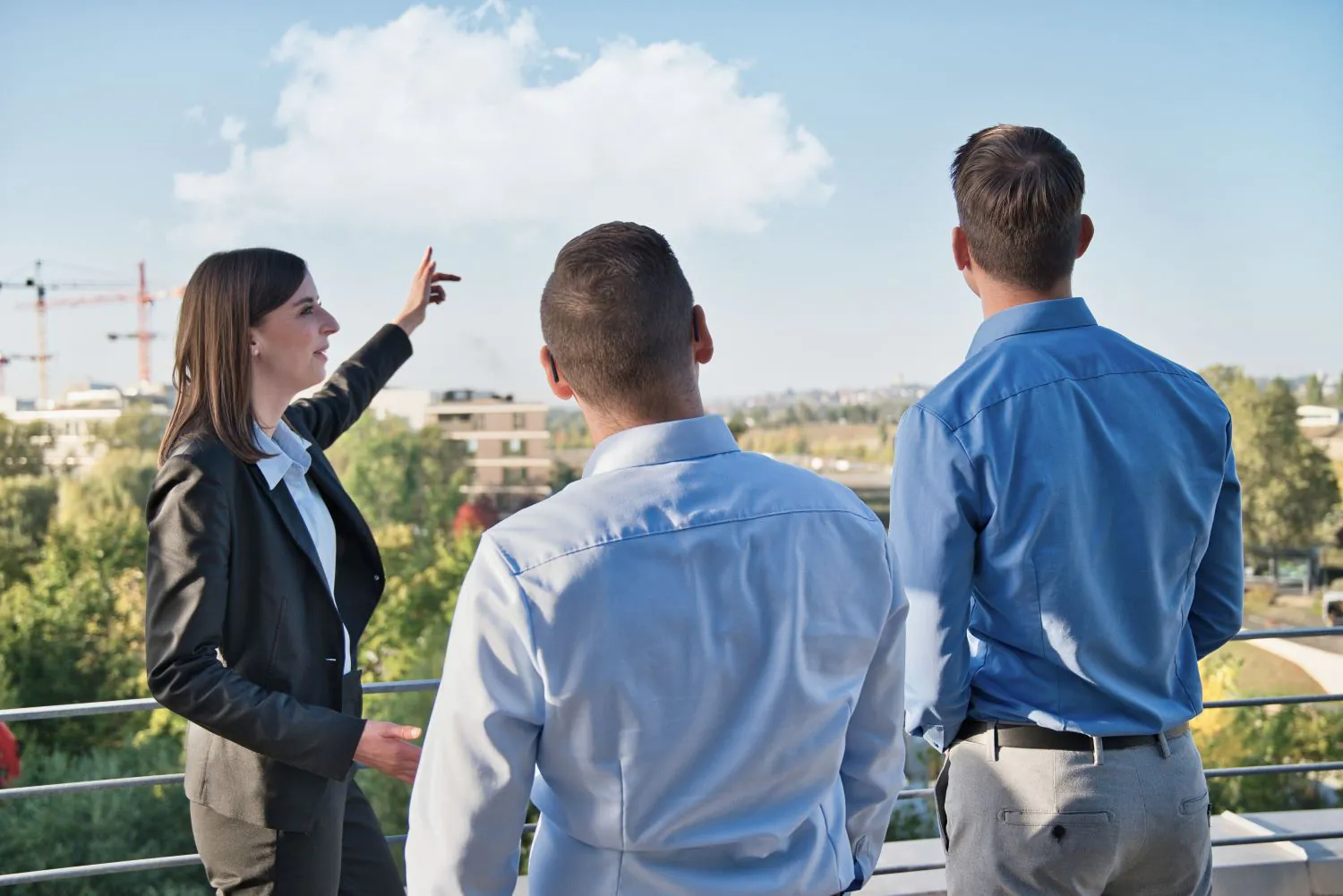 Rückansicht von drei Mitarbeitenden auf einem Balkon die in den blauen Himmel schauen